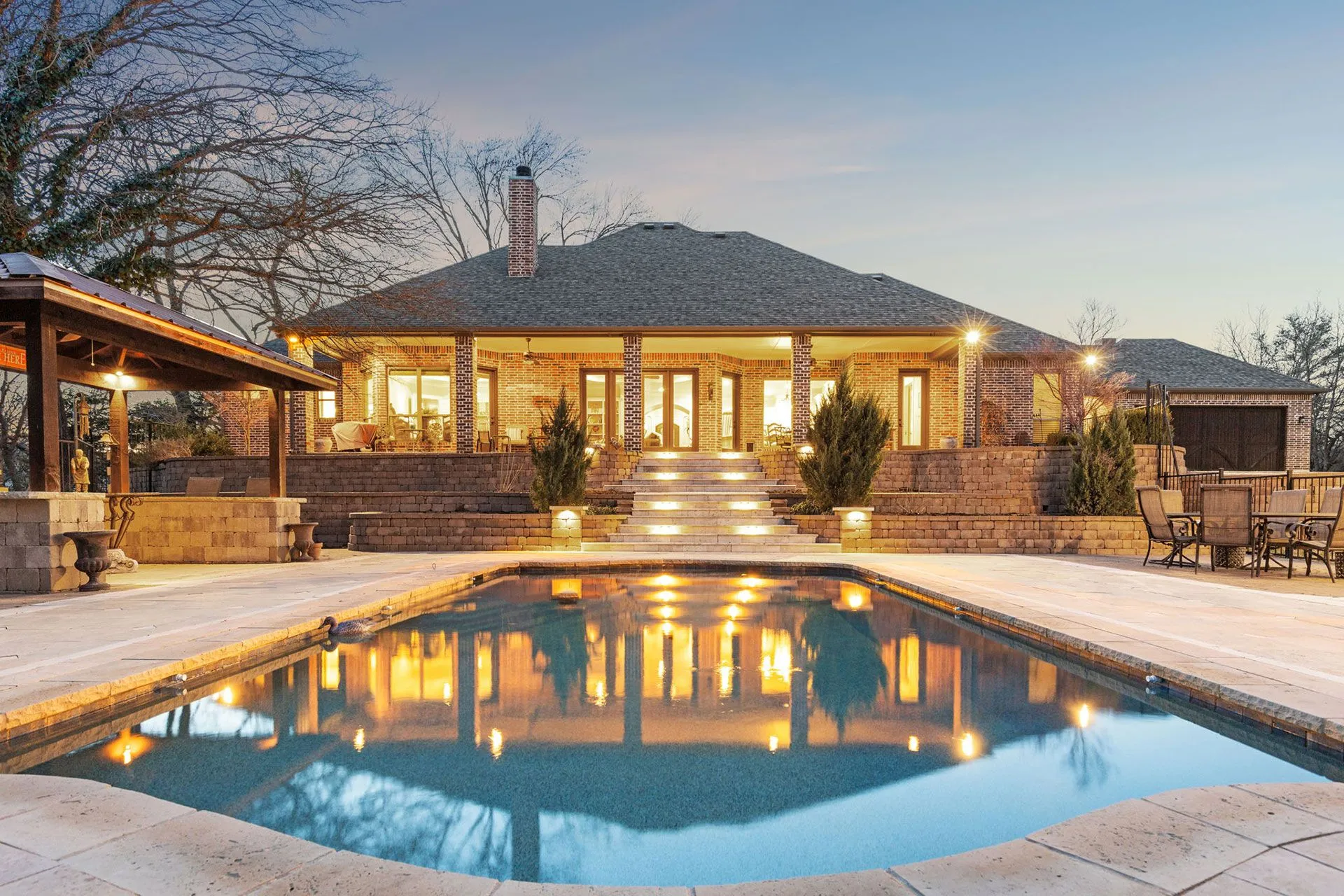 Illuminated pool deck, broad stone steps, and terraced walls behind a North Texas home at twilight
