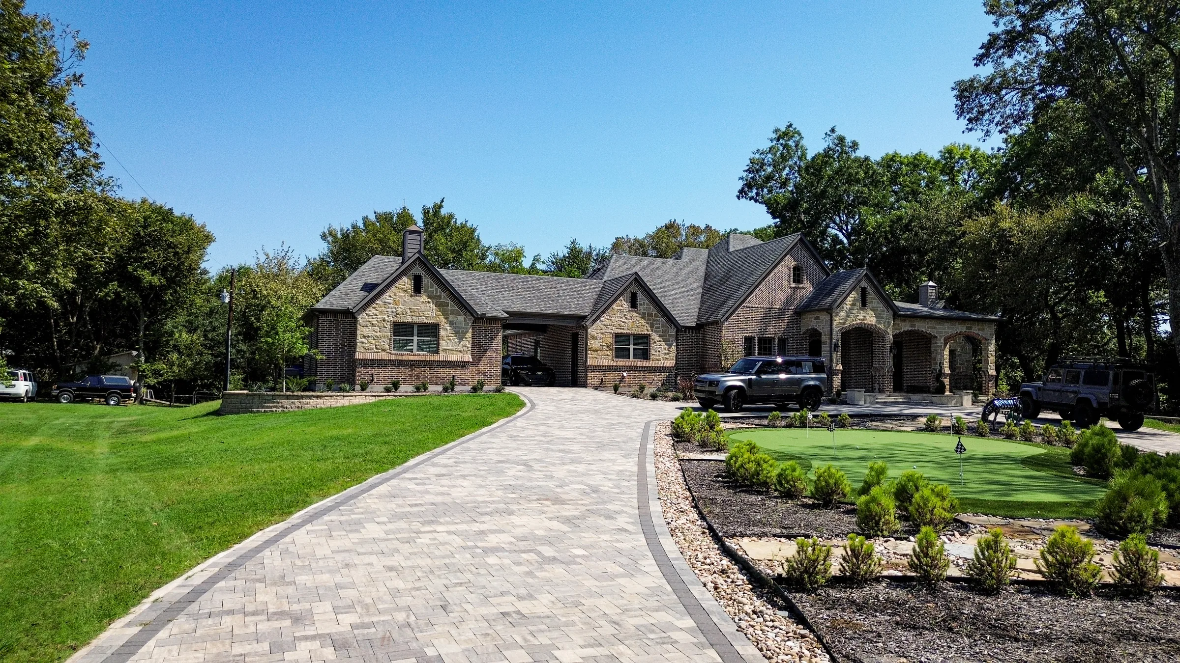 Paver driveway curving toward a stone and brick estate beside a front-yard putting green